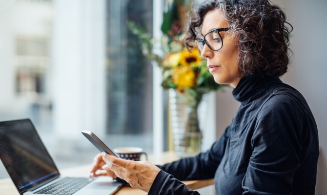Frau vor einem Laptop schaut auf ein Mobiltelefon